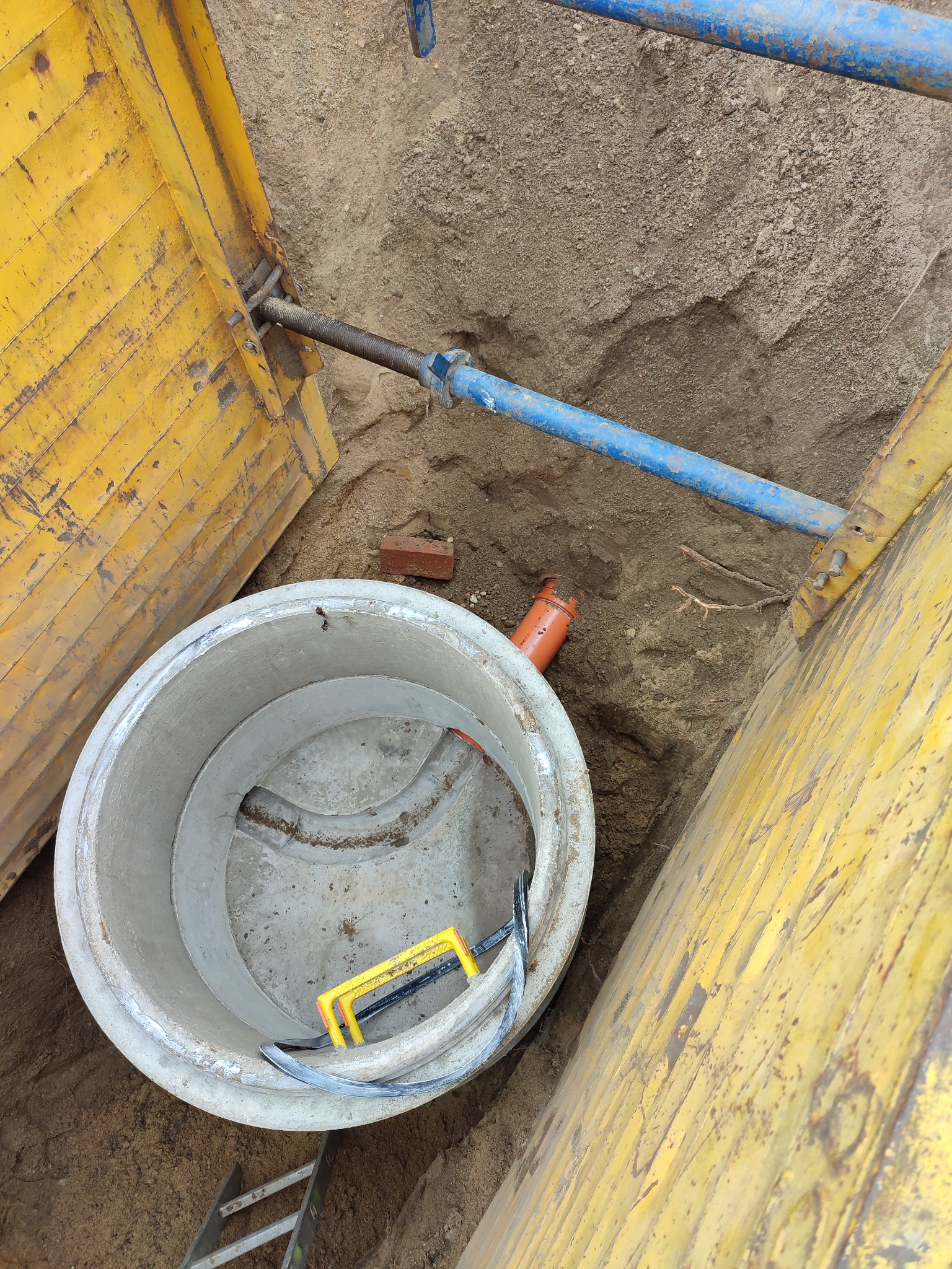 Betonowy krąg studzienny w wykopie zabezpieczonym żółtymi szalunkami, widoczne niebieskie rozpory i fragment pomarańczowej rury.