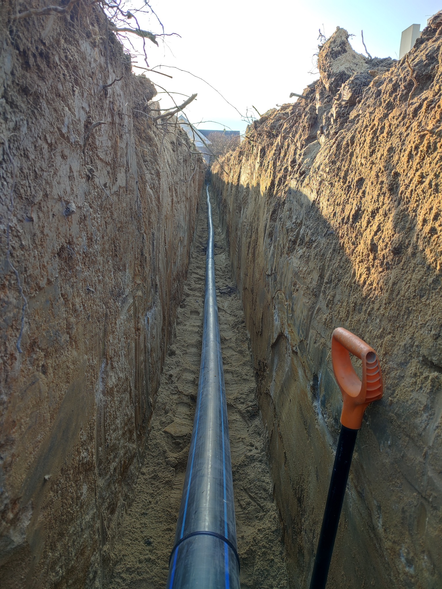 Rów z ułożoną rurą wodociągową z niebieskim oznaczeniem, widoczna pomarańczowa łopata oparta o ścianę wykopu, pionowe ujęcie.