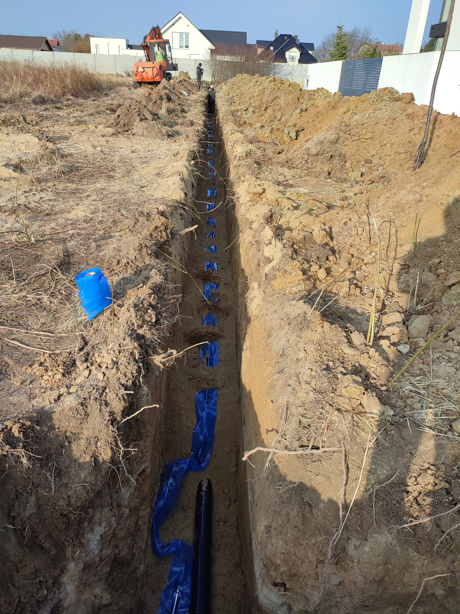 Wykopany rów w ziemi z ułożonymi rurami w niebieskiej folii ochronnej, w tle koparka i zabudowa jednorodzinna.