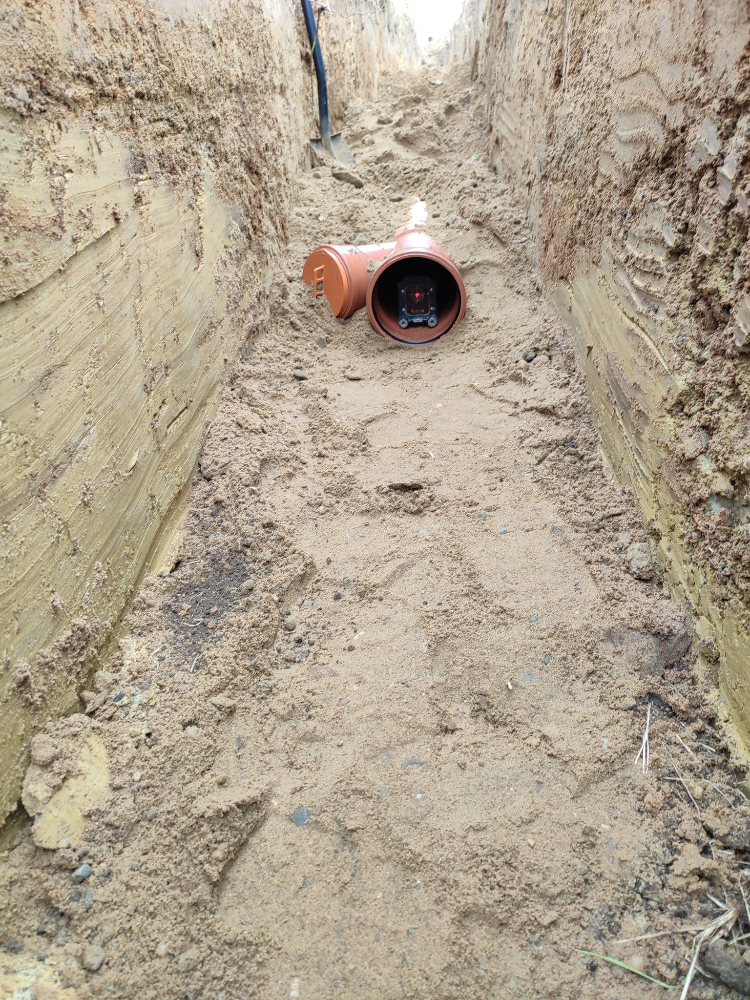 Pomarańczowa rura kanalizacyjna z laserowym niwelatorem umieszczona w wykopie z piaskową podsypką; w tle widoczna łopata.