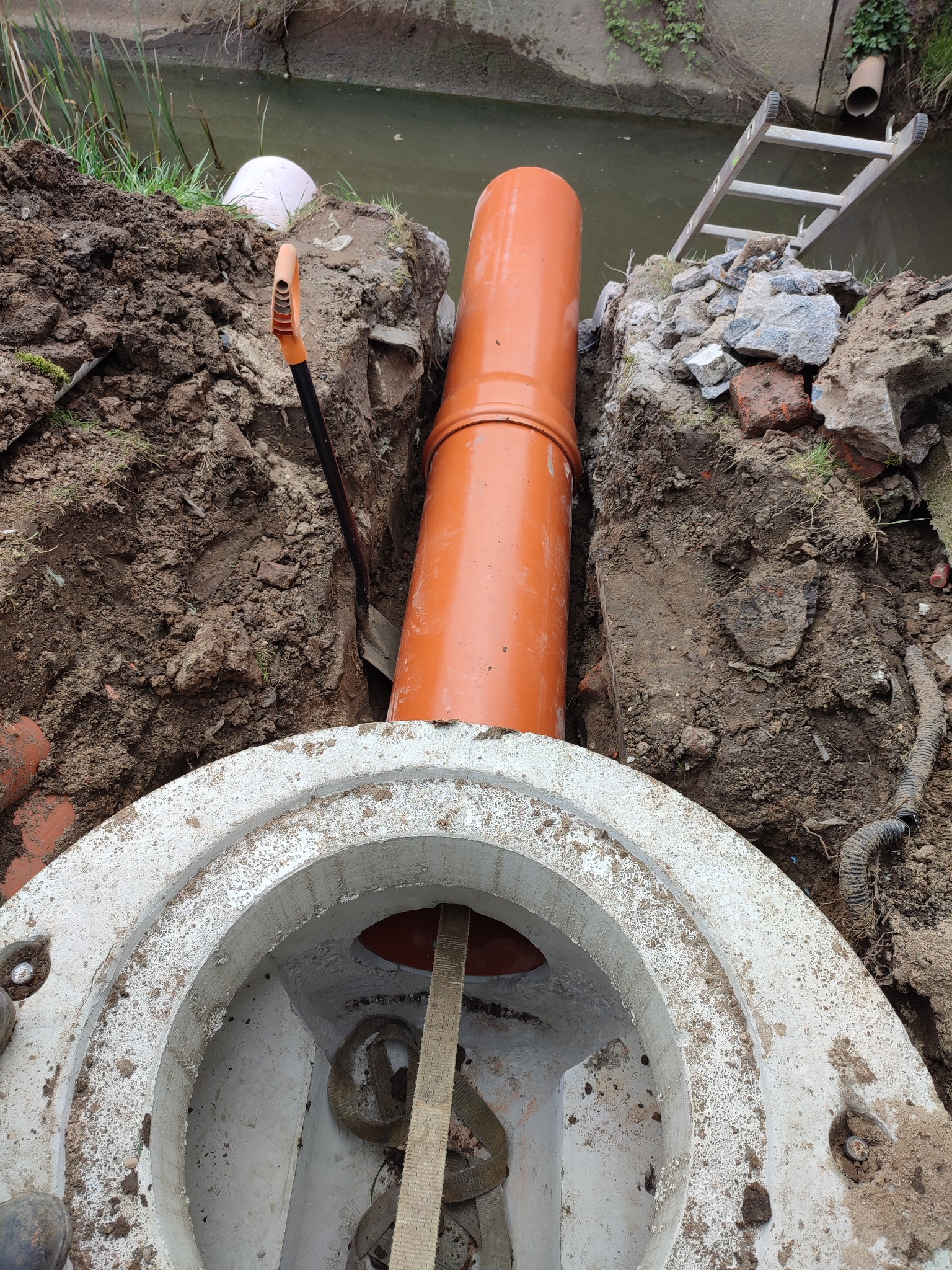 Wykop z pomarańczową rurą kanalizacyjną i betonowym kręgiem studni, widoczna łopata, drabina i brzeg kanału.