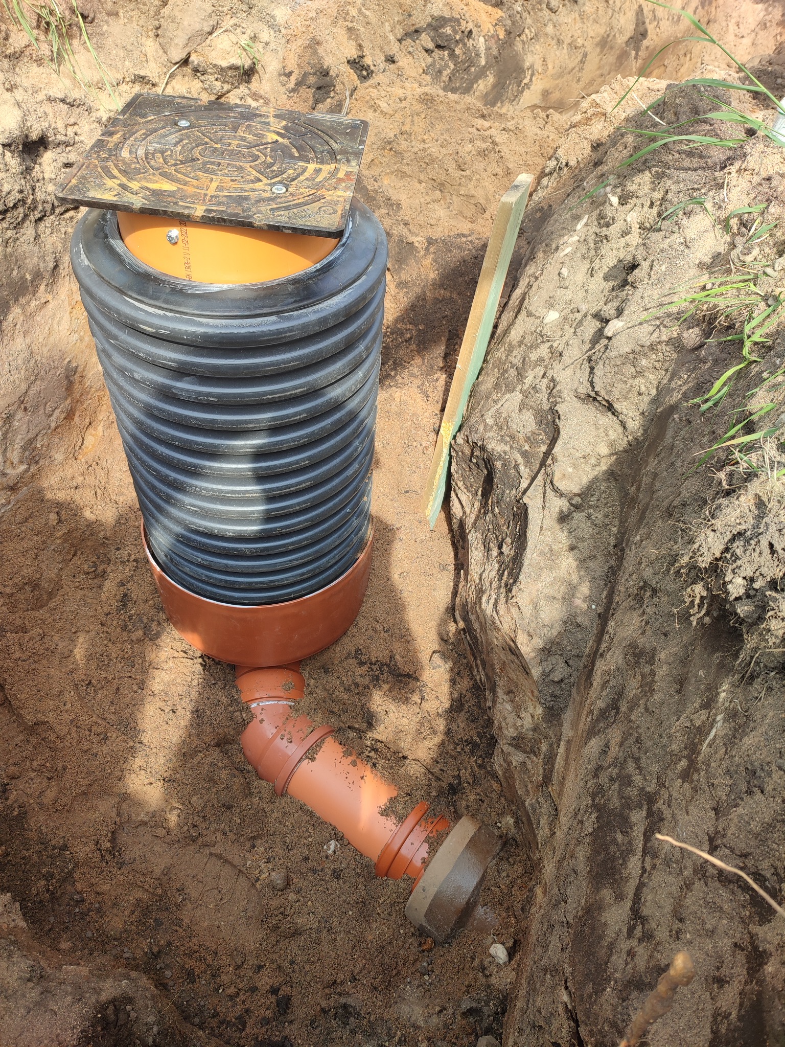 Instalacja zbiornika z rurami w wykopie, widoczna czarna karbowana rura, pomarańczowe elementy i pokrywa włazu na górze, ziemia wokół wykopu.