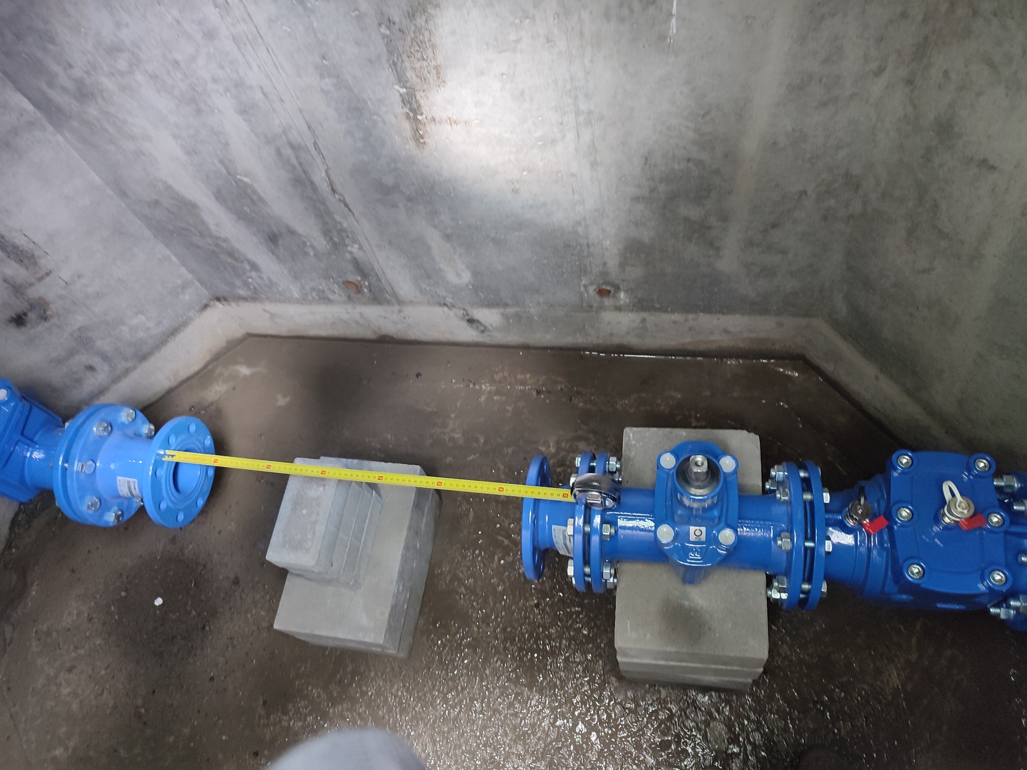 Niebieska rura z zaworami, podparta betonowymi blokami, mierzona żółtą taśmą mierniczą w betonowym kanale