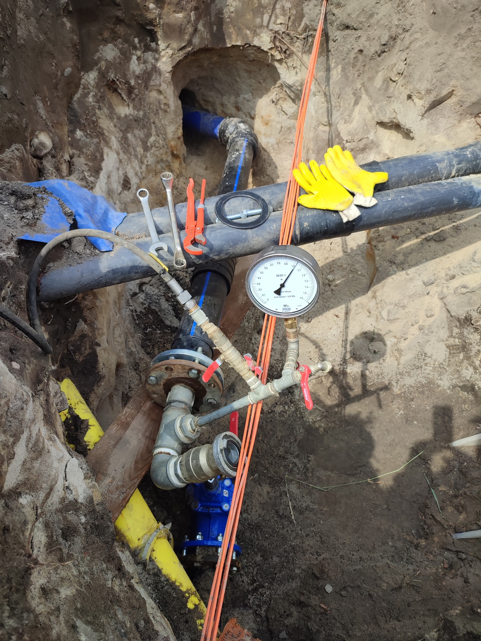 Instalacja wodociągowa w wykopie z widocznym manometrem, żółtymi rękawicami roboczymi, kluczami i fragmentem niebieskiej rury w tle.