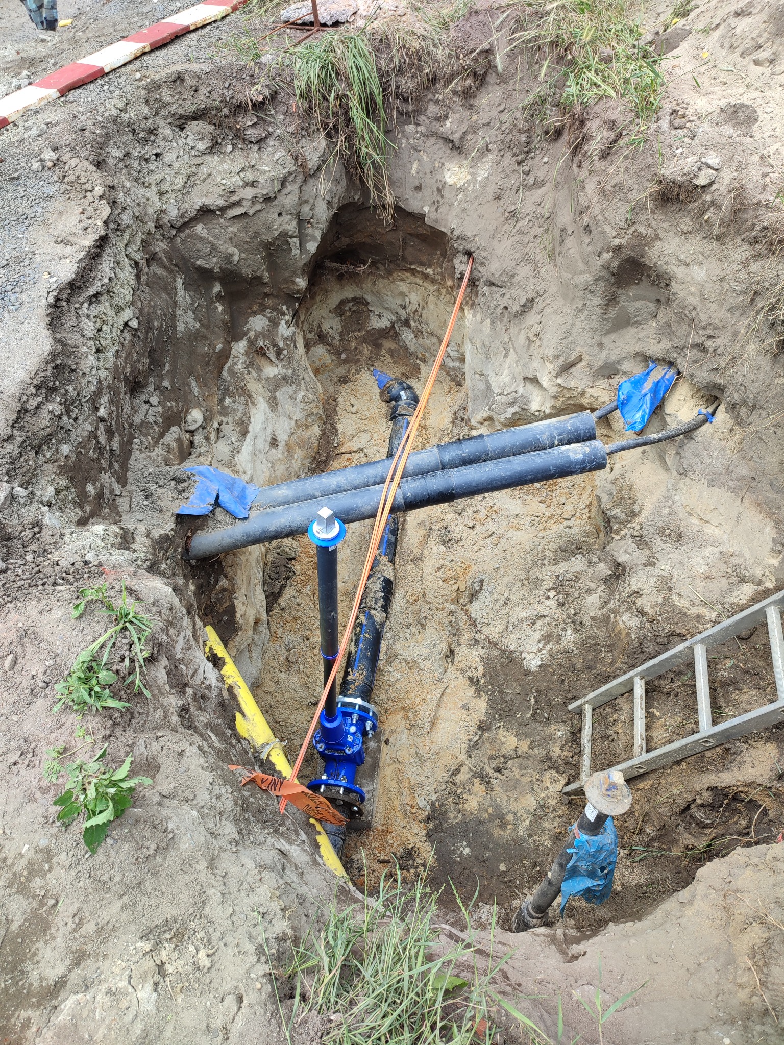 Głęboki wykop w ziemi z widocznymi rurami wodociągowymi, żółtym rurociągiem gazowym, elementami instalacji z niebieskimi kołnierzami i zabezpieczeniami, oraz drabina oparta o krawędź wykopu.