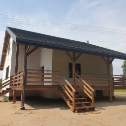 Nowoczesny dom z drewnianym tarasem na podwyższeniu, widoczne schody i balustrady, jasna elewacja, czarny dach, w tle pole i drzewa.