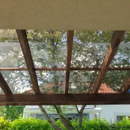 Widok z dołu na drewnianą pergolę z przezroczystym zadaszeniem w Karczewie, przez którą widać drzewa i budynek w tle.