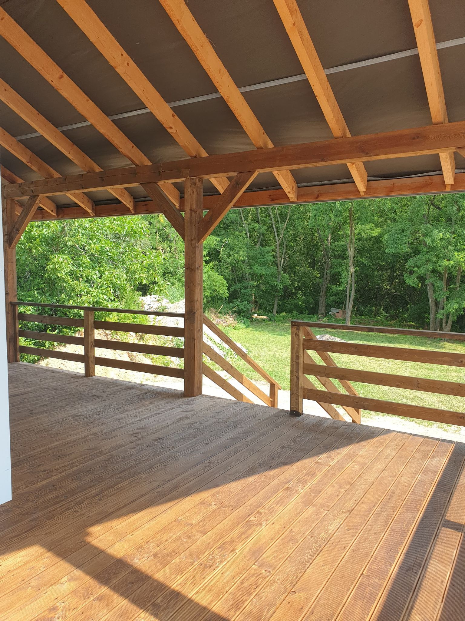 Zadaszony taras z drewna z widokiem na zielony ogród, balustrada z drewnianych belek, naturalne oświetlenie.