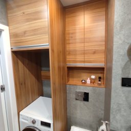 Bez barier - Drewniana zabudowa w łazience z pralką, szafkami wiszącymi i półką z flakonami perfum nad toaletą.