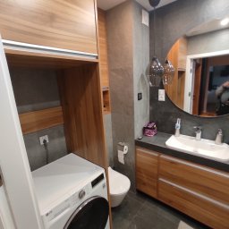 Bez barier - Nowoczesna łazienka z drewnianymi szafkami, umywalką z okrągłym lustrem i pralką w zabudowie z drewnianymi półkami.