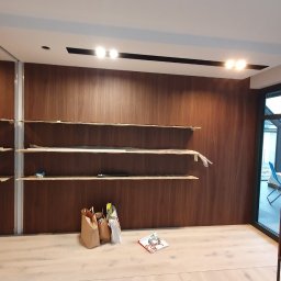 Bez barier - Montaż drewnianej szafy na wymiar z półkami, widoczne elementy konstrukcyjne i materiały budowlane, wnętrze w trakcie wykańczania, okno z widokiem na zewnątrz.