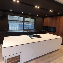 Bez barier - Nowoczesna kuchnia z wyspą: biała wyspa kuchenna z płytą indukcyjną, czarne szafki pod oknem, drewniane szafki po prawej stronie, ściana z tapetą w stylu miejskim, czarny sufit z oświetleniem...