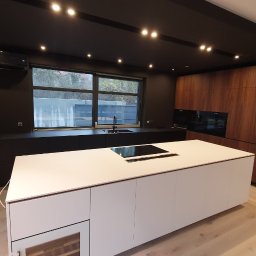 Bez barier - Nowoczesna kuchnia z wyspą, czarnymi szafkami pod oknem, drewnianymi frontami i designerską tapetą w tle. Widoczne oświetlenie punktowe i wbudowany sprzęt AGD.