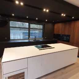 Nowoczesna kuchnia z wyspą, czarnymi szafkami pod oknem, drewnianymi frontami i designerską tapetą w tle. Widoczne oświetlenie punktowe i wbudowany sprzęt AGD.