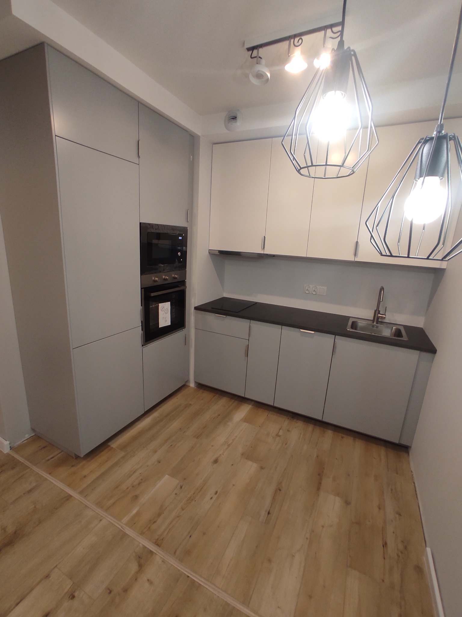 Szara, nowoczesna zabudowa kuchenna z wbudowanym piekarnikiem i mikrofalówką, czarny blat, drewniana podłoga i geometryczne lampy wiszące.