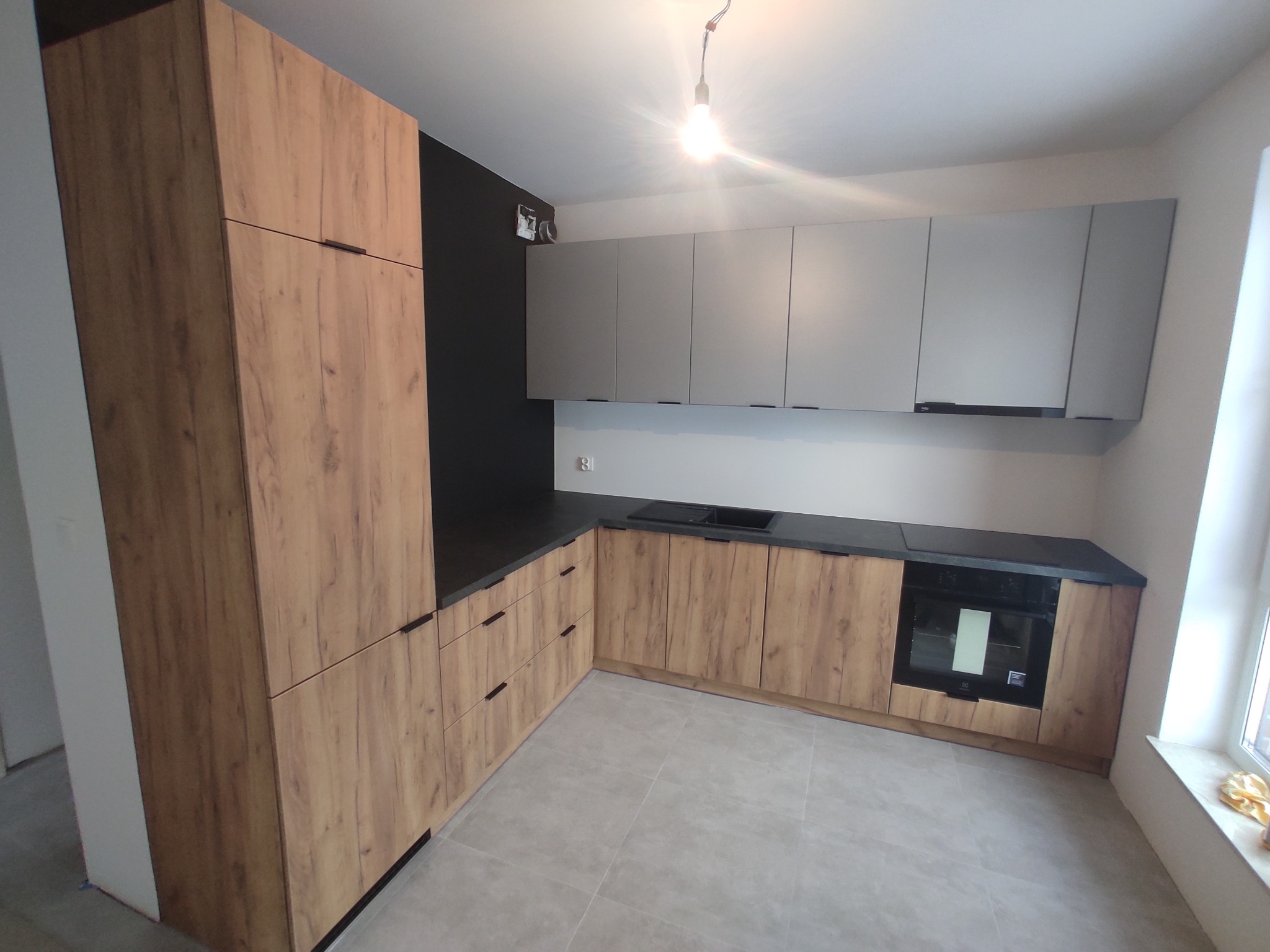 Nowoczesna kuchnia z frontami z drewna o wyraźnym usłojeniu, szare górne szafki i czarny blat, oświetlona żarówką bez klosza.