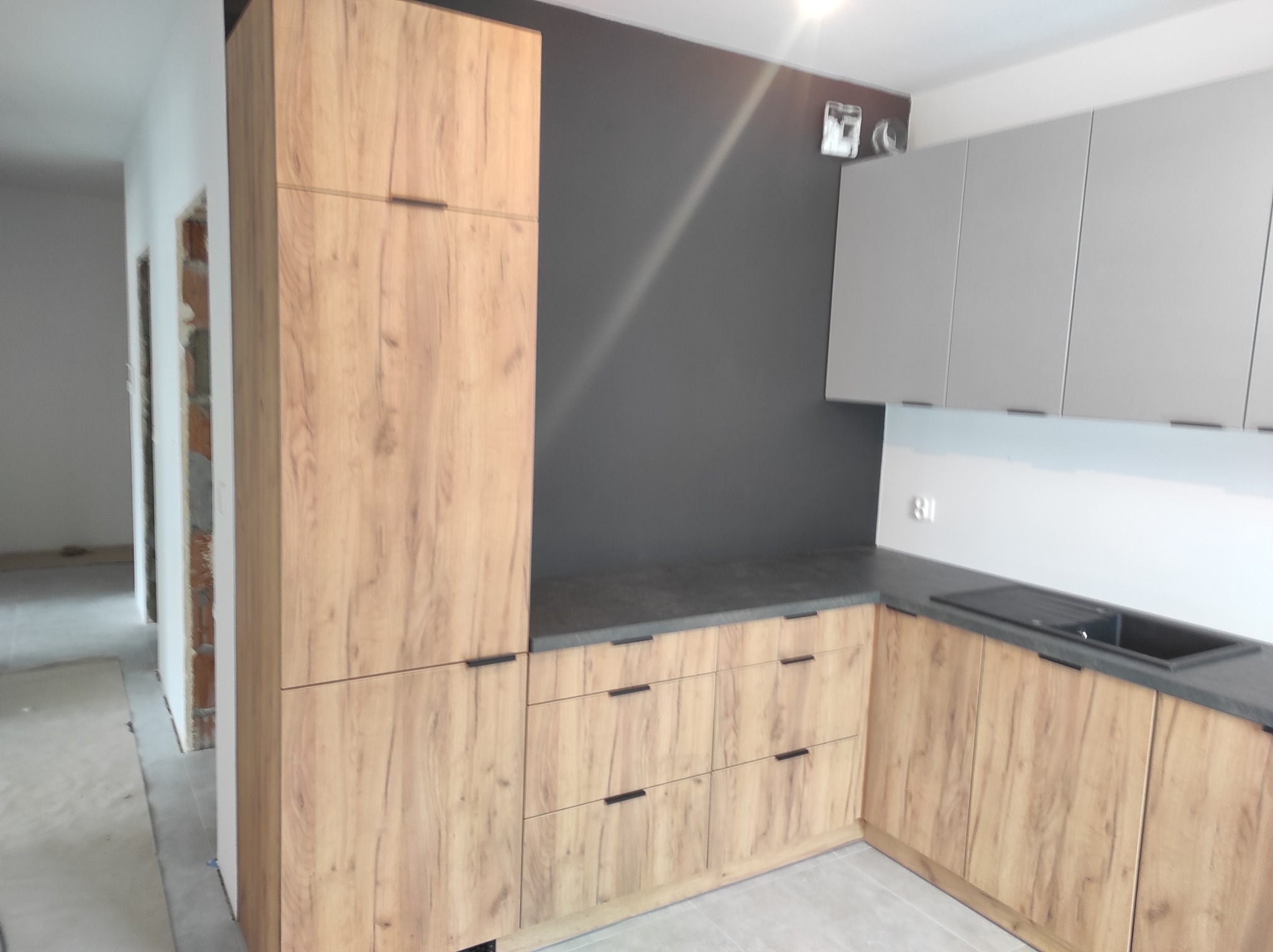 Fragment kuchni z drewnopodobnymi frontami, ciemnoszarym blatem i szarymi szafkami wiszącymi, widoczne niedokończone wykończenie ścian w tle.