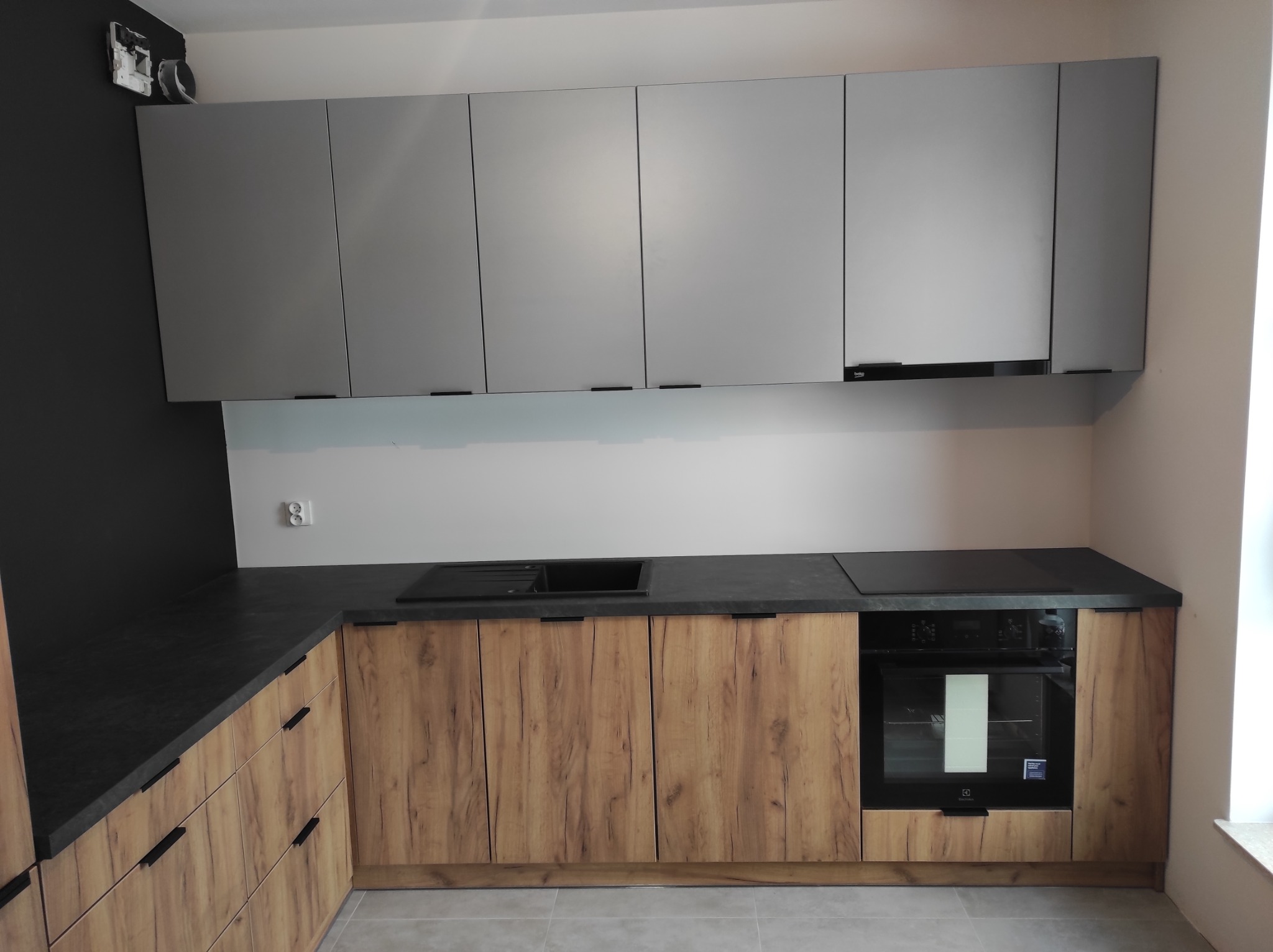 Nowoczesna kuchnia z szarymi szafkami górnymi i drewnopodobnymi szafkami dolnymi, czarny blat, płyta indukcyjna i piekarnik.