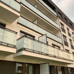 Nowoczesny budynek wielorodzinny z balkonami zabezpieczonymi szklanymi balustradami z czarnymi ramami, widok z dołu na kilka pięter.
