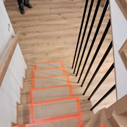 STOLARSTOW MARIUSZ BOCIAN - Widok z góry na drewniane schody w trakcie montażu, zabezpieczone folią ochronną z pomarańczową taśmą, z czarnymi, metalowymi balustradami.