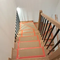 Klatka schodowa w trakcie zabezpieczania, drewniane stopnie oklejone kartonem i pomarańczową taśmą, czarne metalowe barierki, widok z góry.