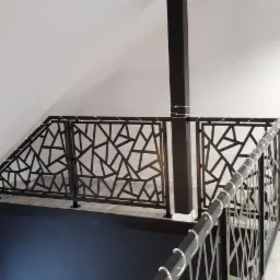 Czarna, metalowa balustrada antresoli z geometrycznym wzorem, widok z góry, z elementami dekoracyjnymi z przezroczystego akrylu na poręczy. W tle widoczny filar i fragment pomieszczenia z telewizorem.