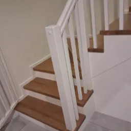 Fragment drewnianych schodów z białymi barierkami, widok z dołu na dwa stopnie z szarej podłogi.