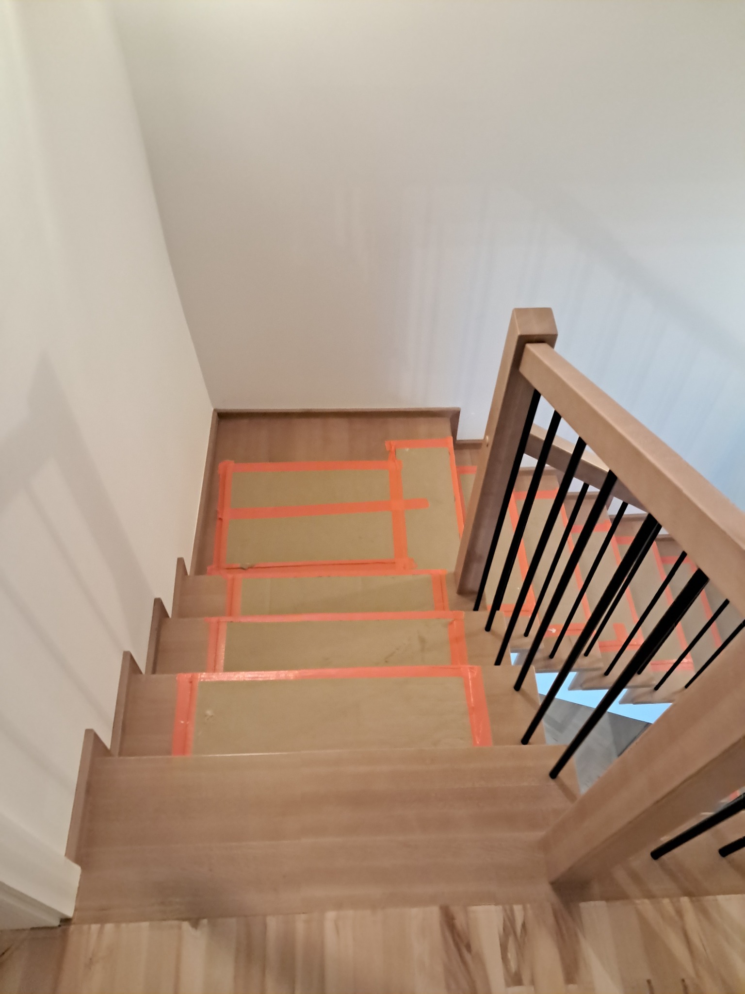 Wnętrze domu w trakcie wykańczania: widok z góry na drewniane schody z zabezpieczonymi stopniami kartonem i taśmą malarską, z jasną drewnianą balustradą z czarnymi, pionowymi elementami.