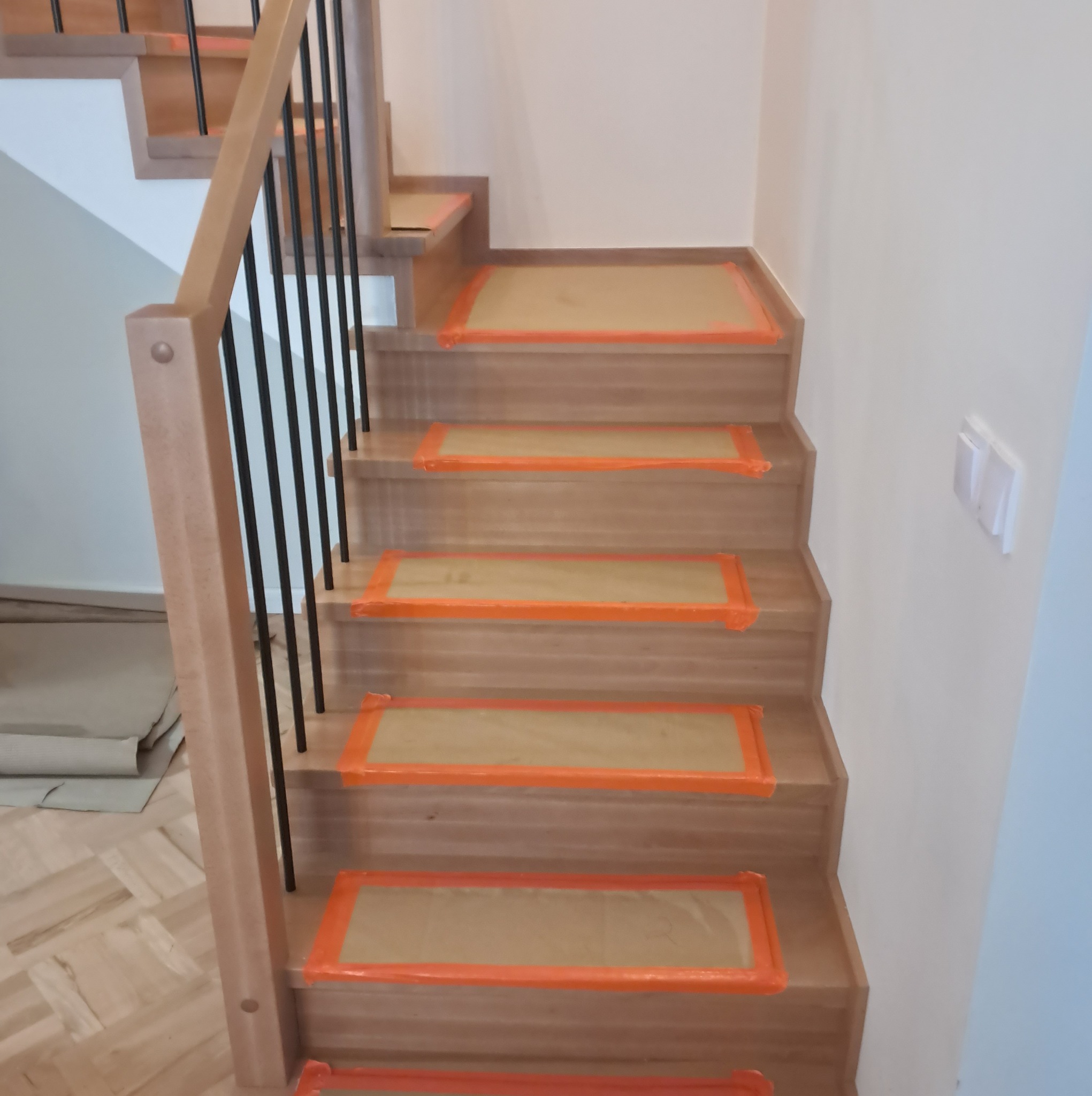 Jasne drewniane schody z czarnymi, metalowymi balustradami, zabezpieczone folią ochronną na stopniach, widok z dołu.