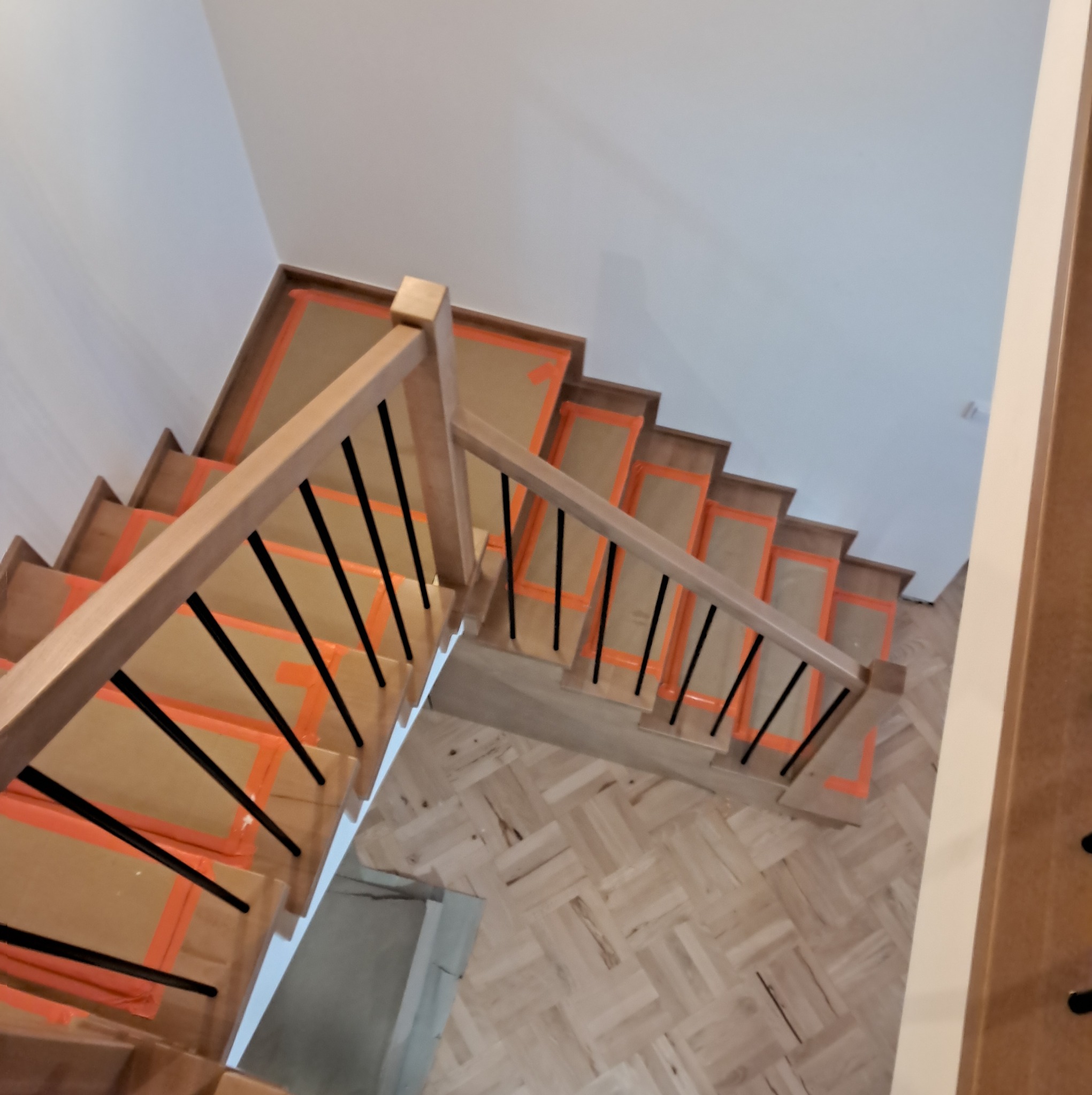 Kątowy widok z góry na drewniane schody z czarnymi balustradami, zabezpieczone kartonem i pomarańczową taśmą malarską, prowadzące na parkiet ułożony w jodełkę.