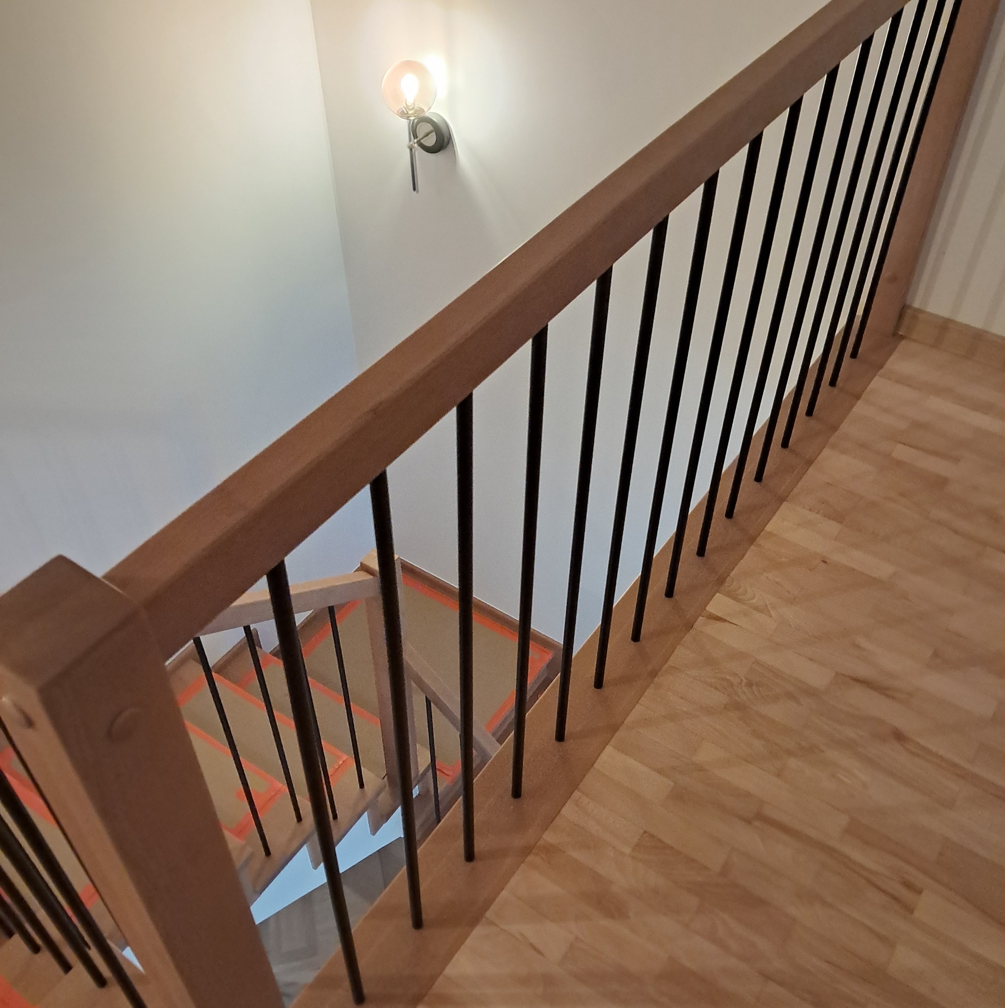 Nowoczesne schody z drewnianymi stopniami zabezpieczonymi pomarańczową taśmą, czarne metalowe balustrady i drewniana poręcz, oświetlone kinkietem z żarówką w stylu retro.