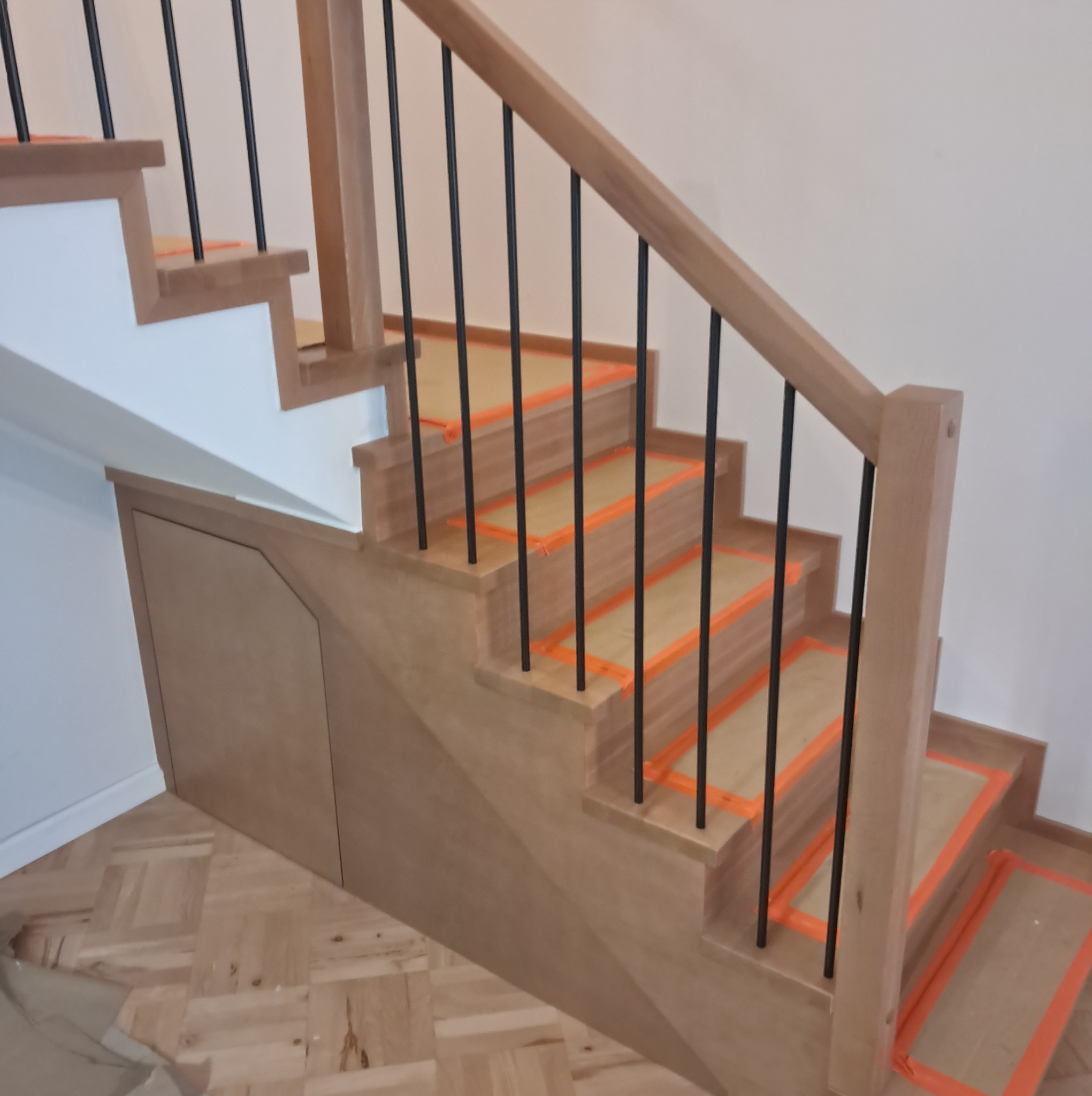Drewniane schody z czarnymi balustradami zabezpieczone pomarańczową taśmą malarską, z zabudowaną szafką pod schodami i parkietem na podłodze.
