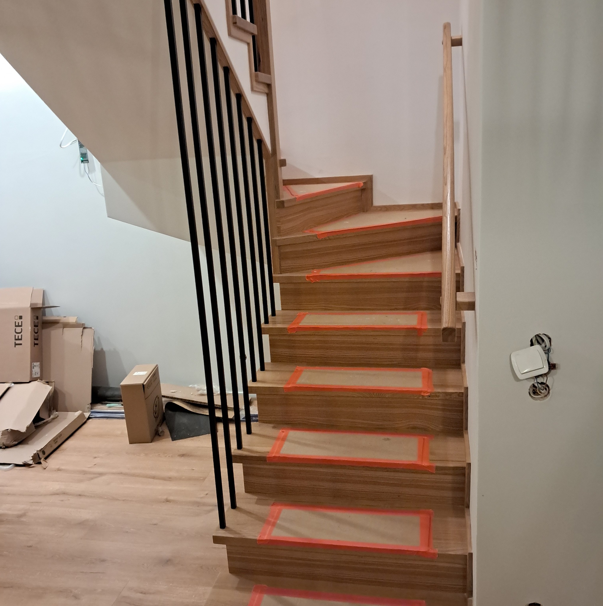 Drewniane schody z czarną balustradą w trakcie montażu, zabezpieczone folią ochronną, widoczne kartony po produktach budowlanych w tle.