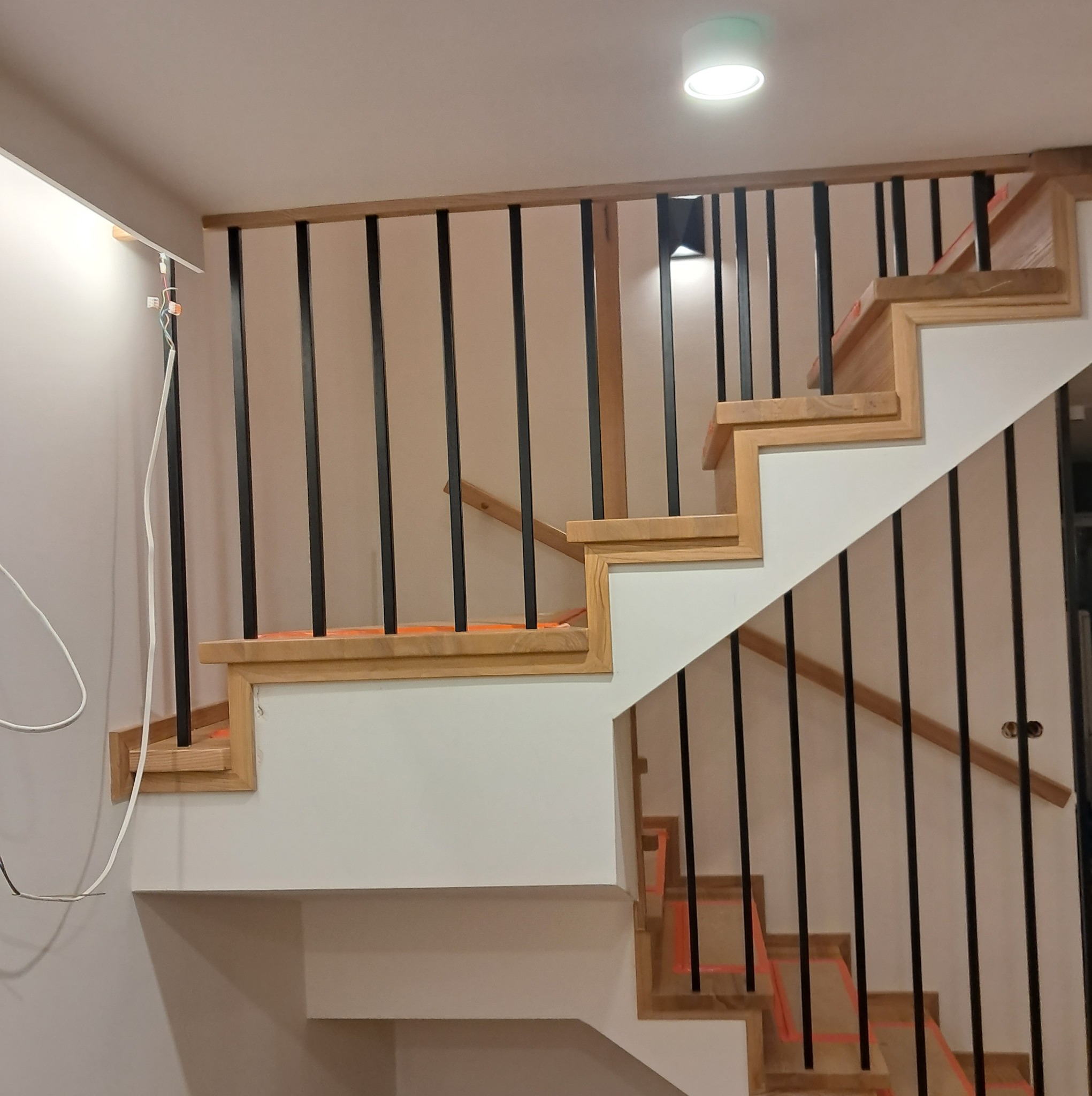 Fragment konstrukcji schodów z drewnianymi stopniami zabezpieczonymi pomarańczową folią, czarne balustrady, białe podstopnice, widoczne przewody elektryczne.
