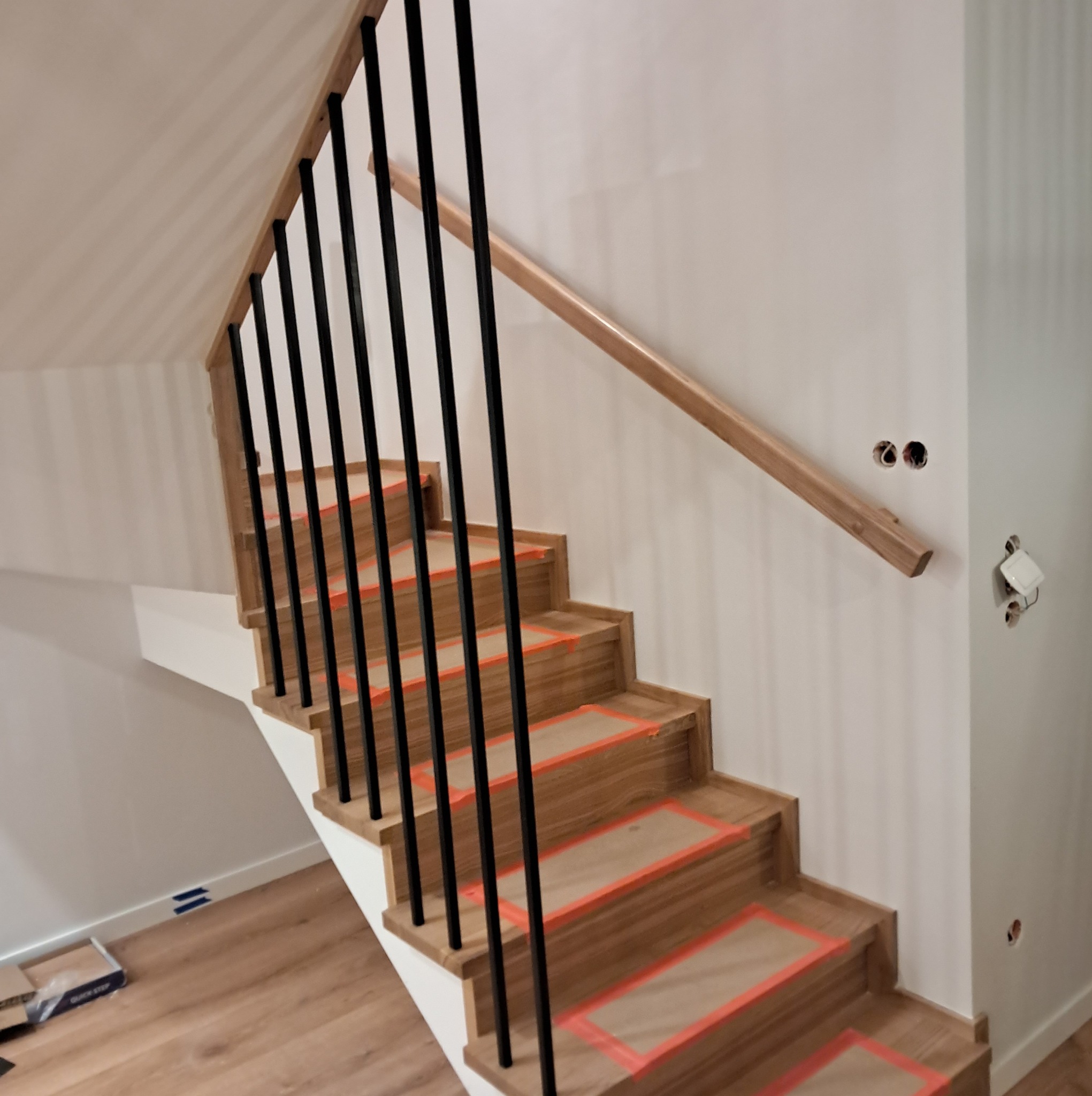 Dębowe stopnie schodów z czarnymi balustradami zabezpieczone kartonem i taśmą malarską, widoczne puszki elektryczne w ścianie i opakowanie paneli podłogowych.