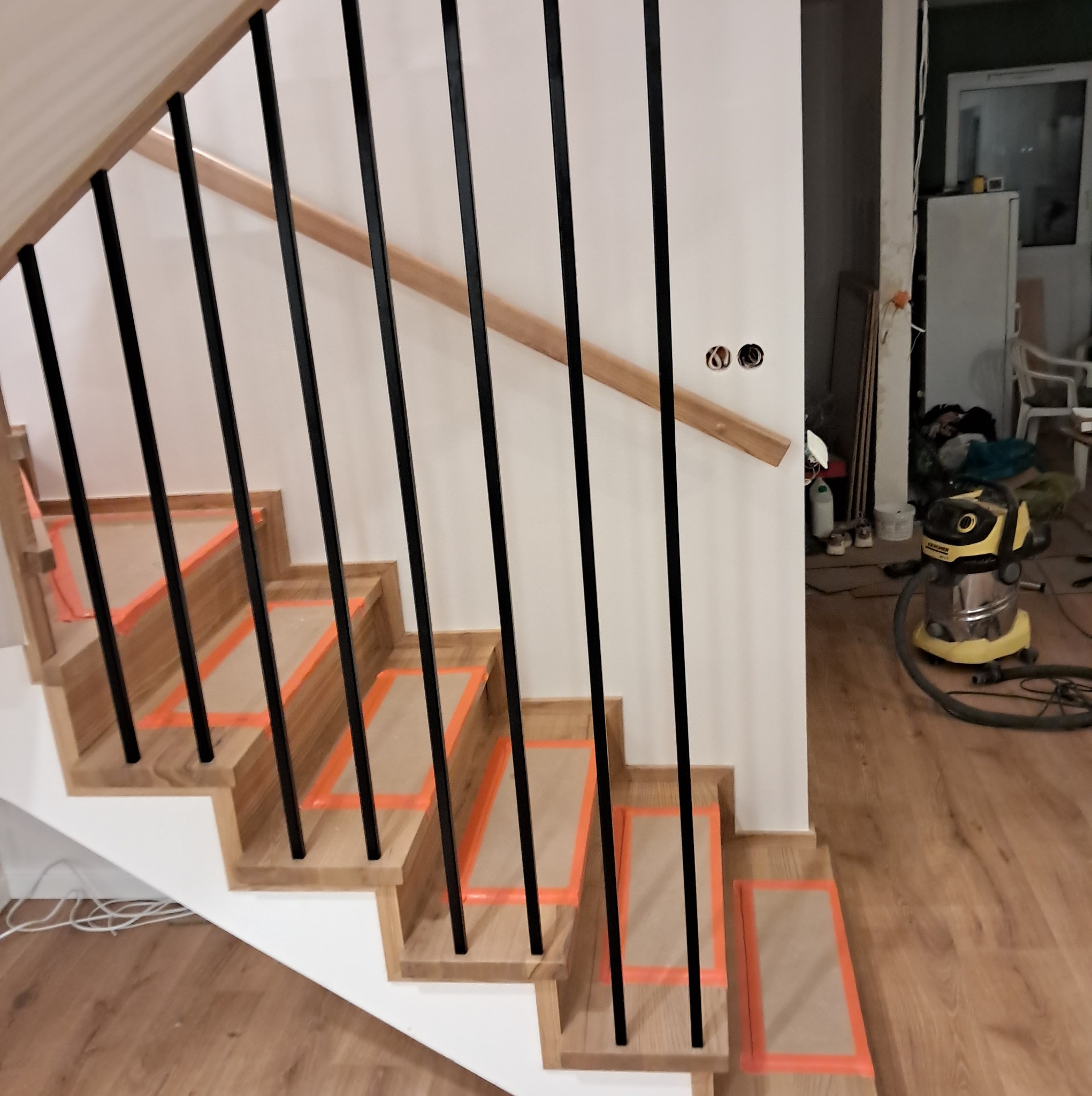 Nowoczesne schody z drewnianymi stopniami zabezpieczonymi pomarańczową taśmą malarską i czarnymi metalowymi balustradami, widoczne podczas montażu, z odkurzaczem przemysłowym w tle.