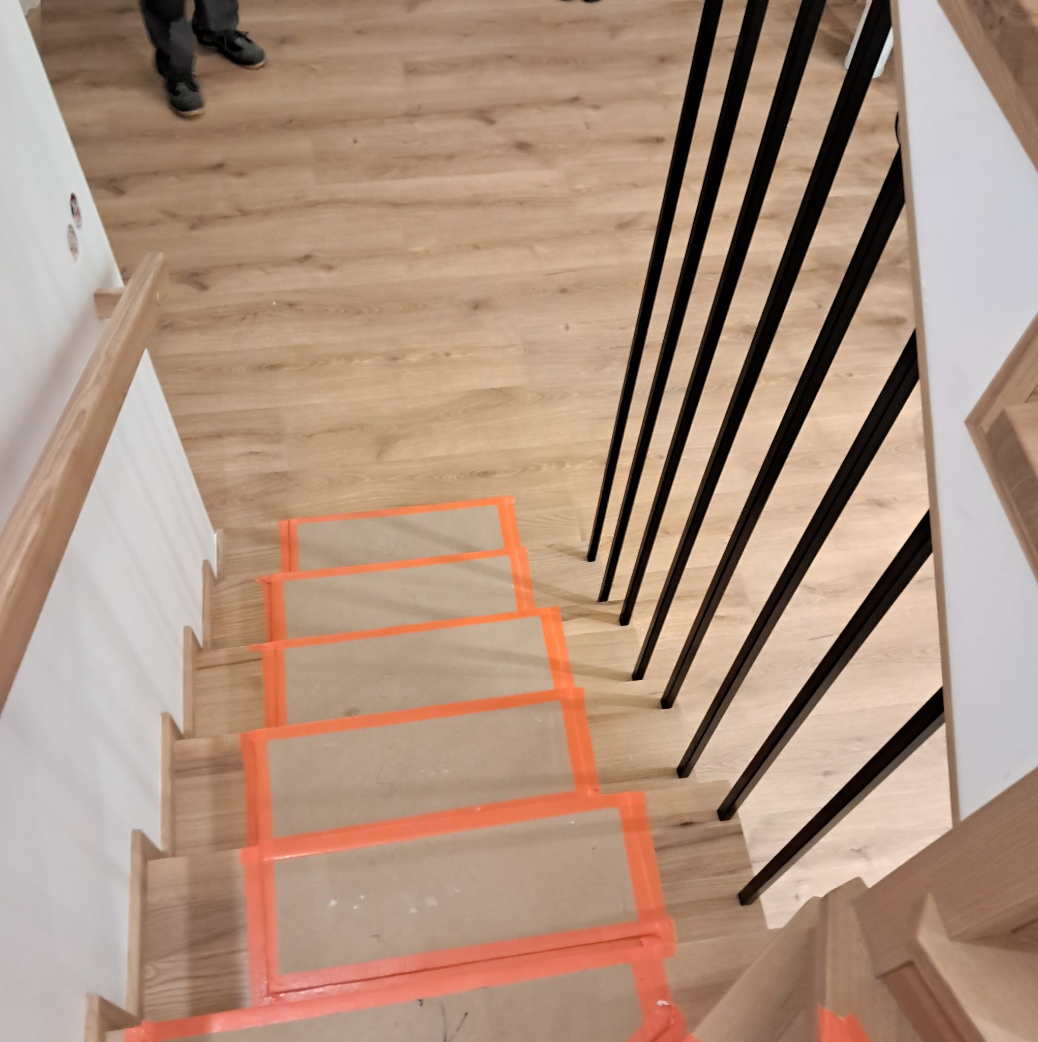 Widok z góry na drewniane schody w trakcie montażu, zabezpieczone folią ochronną z pomarańczową taśmą, z czarnymi, metalowymi balustradami.
