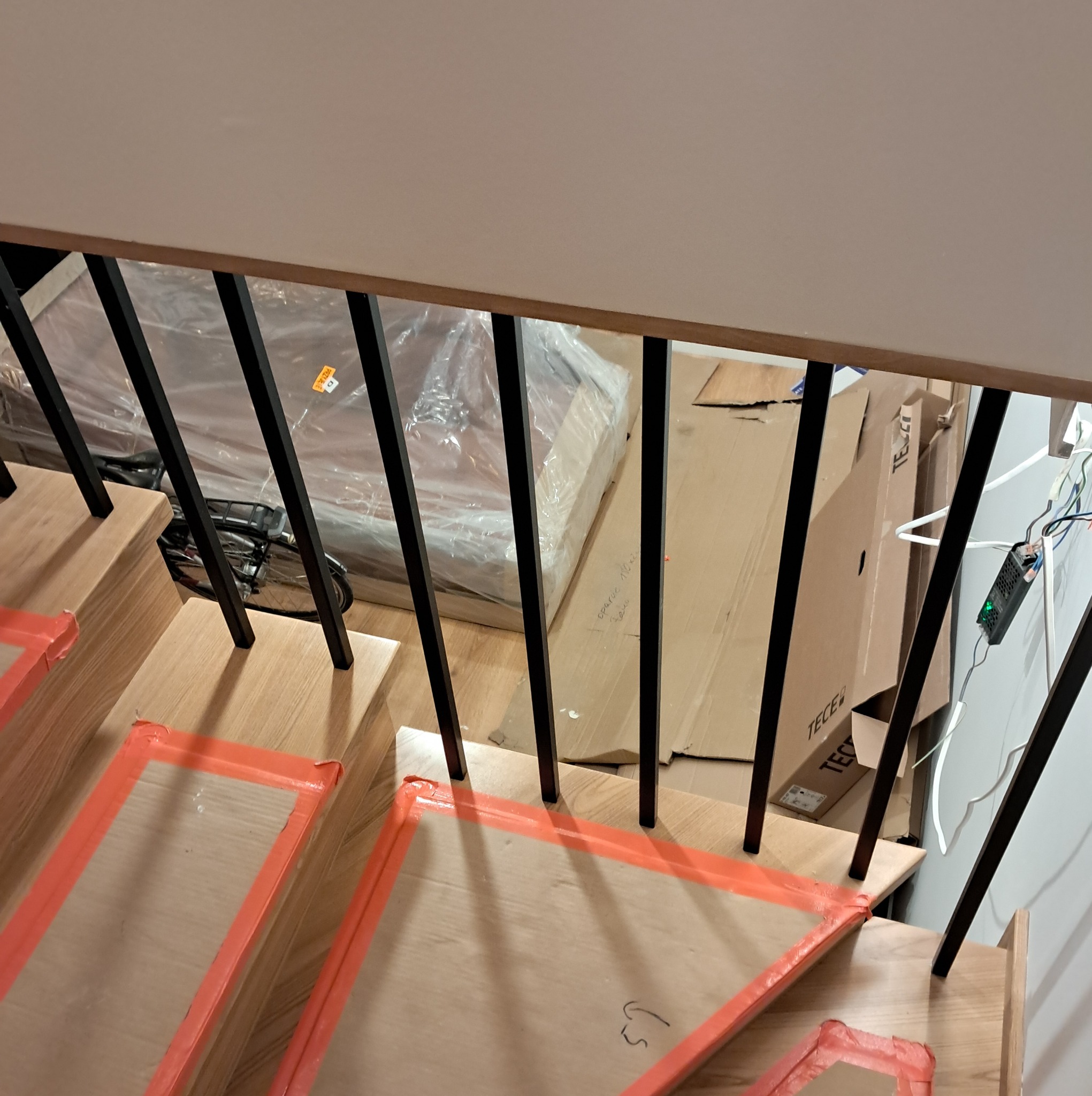 Montaż drewnianych schodów z czarną balustradą, zabezpieczone pomarańczową taśmą, widoczne kartony i rower w tle.