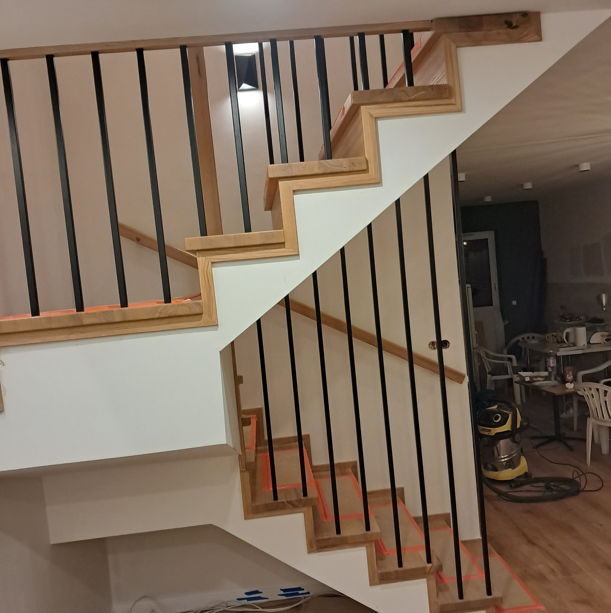 Drewniane schody z czarnymi balustradami w trakcie montażu, zabezpieczone taśmą malarską, widoczny odkurzacz przemysłowy w tle.
