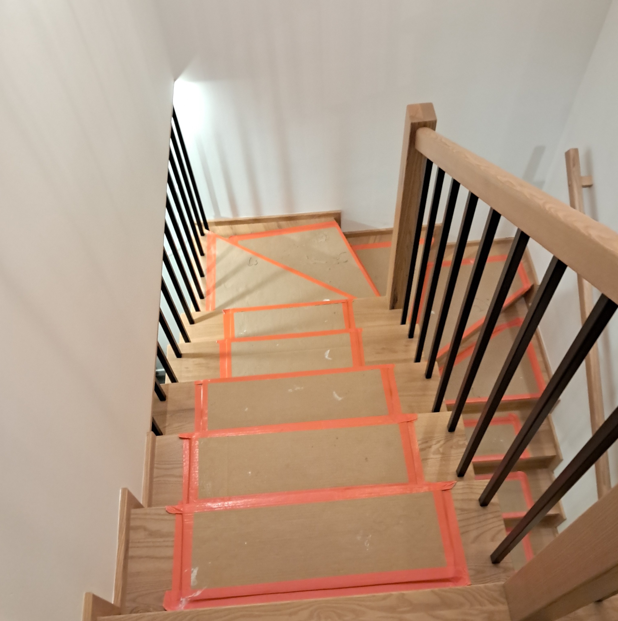 Klatka schodowa w trakcie zabezpieczania, drewniane stopnie oklejone kartonem i pomarańczową taśmą, czarne metalowe barierki, widok z góry.
