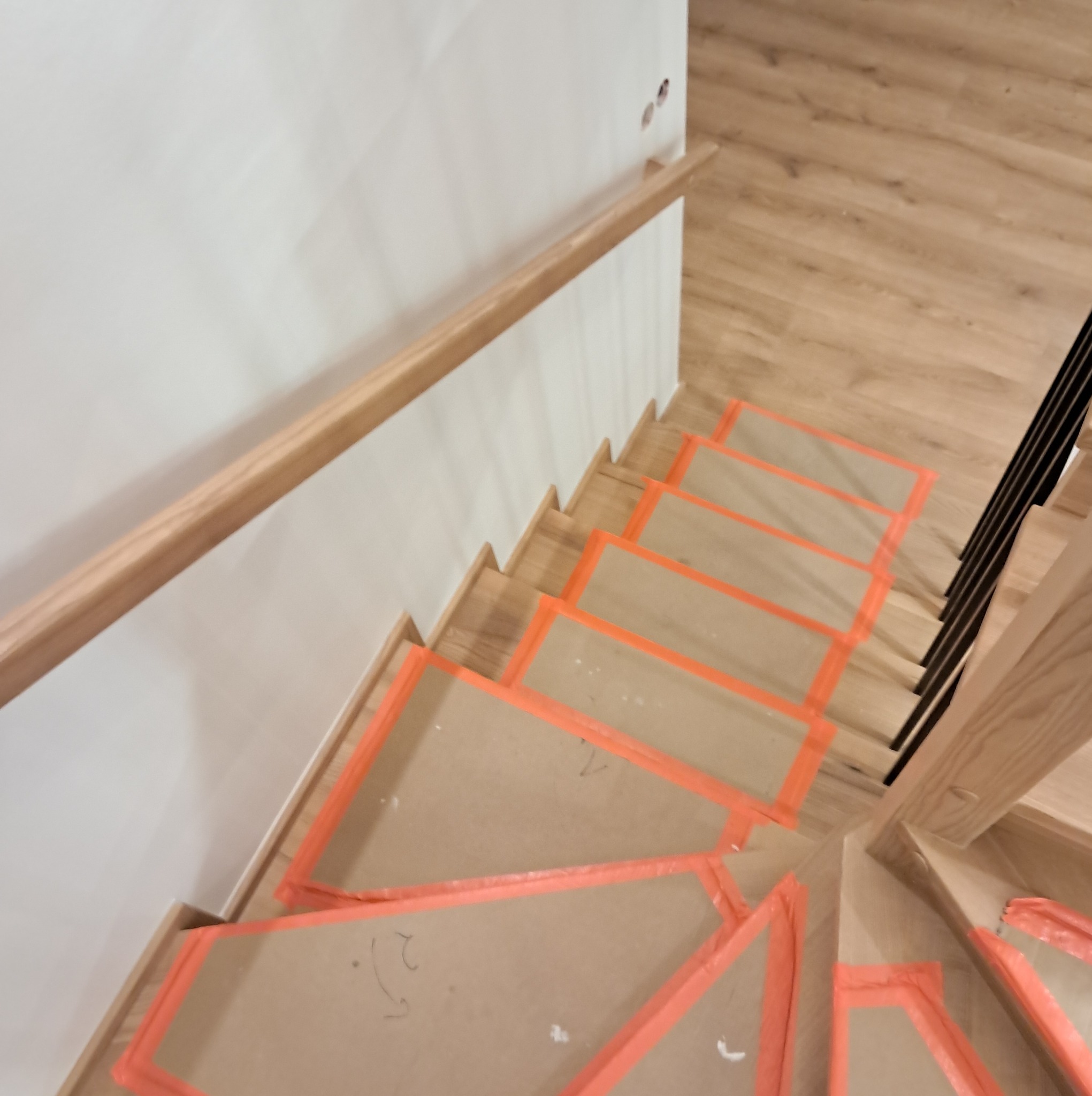 Drewniane schody zabezpieczone kartonem i pomarańczową taśmą malarską podczas remontu, widok z góry na stopnie i fragment balustrady.