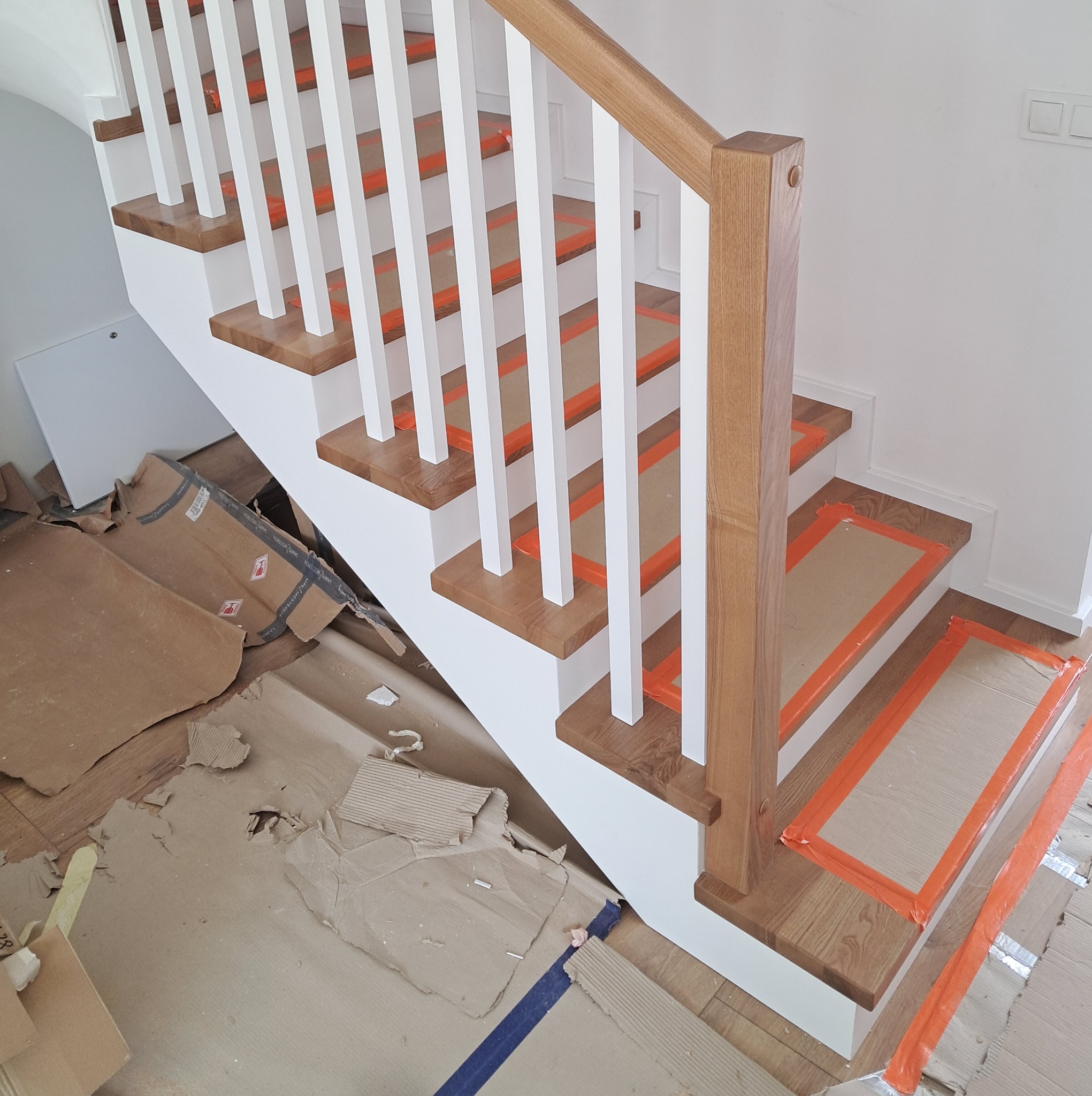 Wnętrze z drewnianymi schodami z białymi balustradami, zabezpieczonymi pomarańczową taśmą malarską, w trakcie montażu lub tuż po nim, z widocznymi kartonami i resztkami materiałów na podłodze.