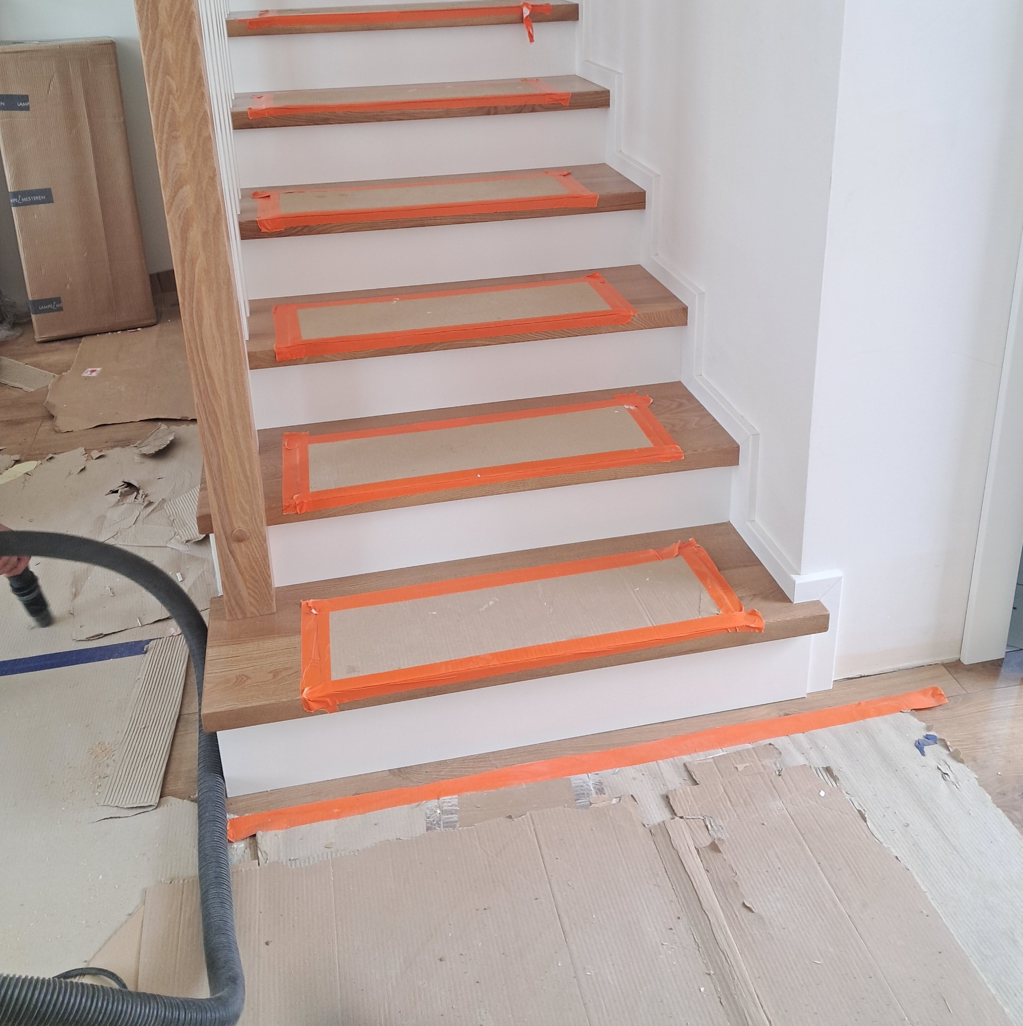 Montaż drewnianych stopni schodów zabezpieczonych pomarańczową taśmą i tekturą, widoczny odkurzacz przemysłowy i kartony na podłodze.