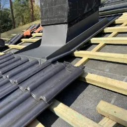 Montaż czarnej dachówki ceramicznej wokół komina z blachy, widoczna drewniana konstrukcja dachu i torba narzędziowa w tle.