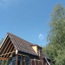 Dom w trakcie budowy, widoczna konstrukcja dachu z łatami i membraną, rusztowania, drabina oparta o ścianę, błękitne niebo w tle.