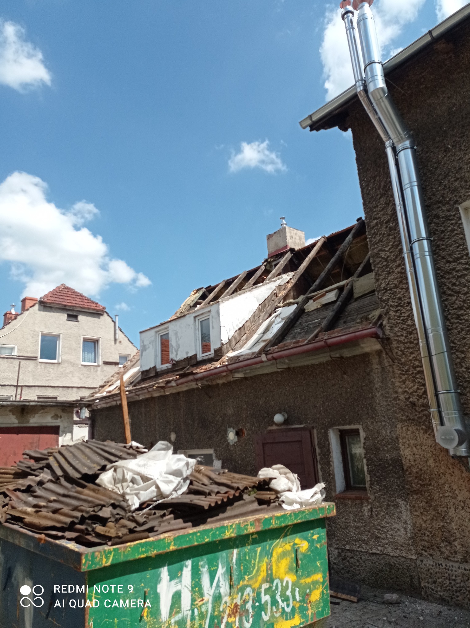 Uszkodzony dach budynku w trakcie demontażu, widoczne fragmenty więźby dachowej, obok kontener ze zdemontowanymi elementami pokrycia i nowa, metalowa instalacja kominowa.