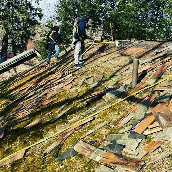 Demontaż starego, zniszczonego dachu z mchem i częściowo usuniętą dachówką przez dwóch dekarzy zabezpieczonych linami, widoczny komin i otoczenie drzew.