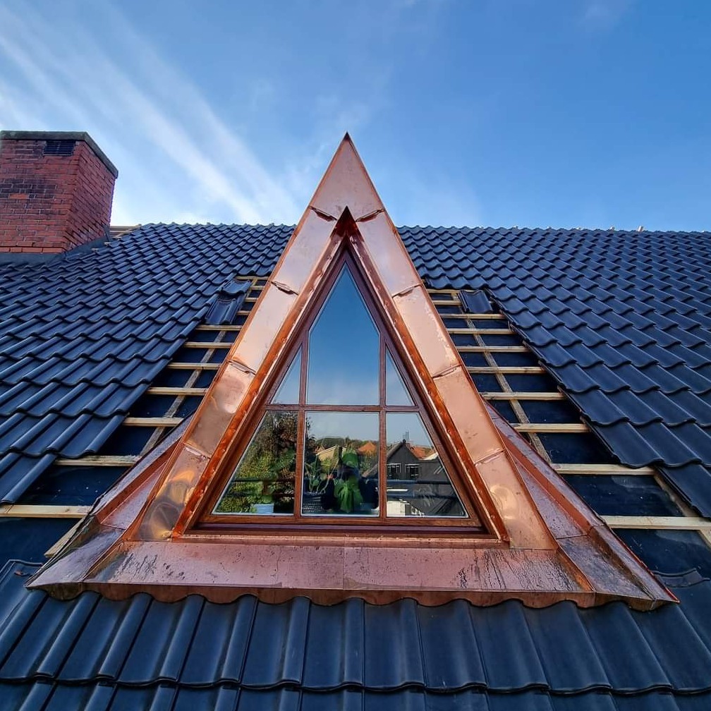 Trójkątne okno dachowe z miedzianą obróbką blacharską, widoczne na tle niebieskiego nieba i częściowo pokrytego czarną dachówką dachu, z fragmentami drewnianej konstrukcji pod spodem.
