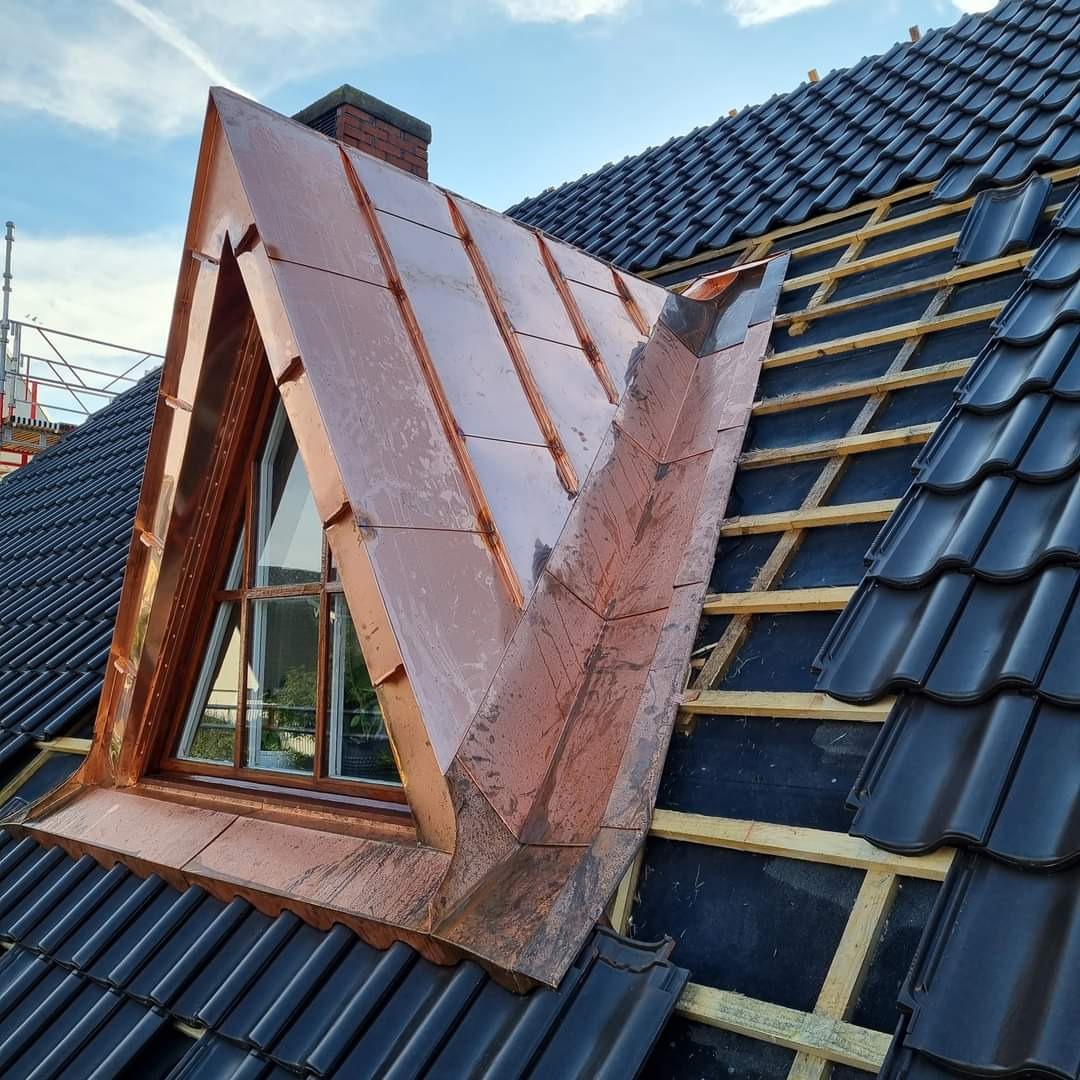 Miedziana obróbka blacharska okna dachowego typu lukarna na tle fragmentu dachu z czarnej dachówki i odsłoniętej więźby. Widoczne ślady po deszczu.