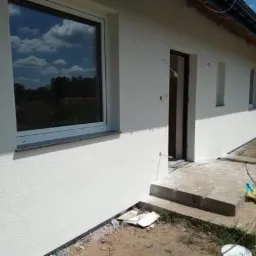 Biała elewacja domu jednorodzinnego z widocznym oknem, drzwiami wejściowymi i fragmentem dachu, teren wokół w trakcie prac wykończeniowych, widoczne zabrudzenia i resztki materiałów budowlanych.